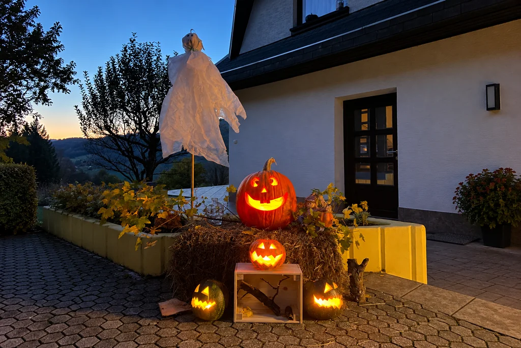 One white fabric ghost above four carved pumpkin lanterns, a hay bale and a wooden crate outside a house